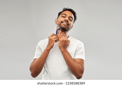 Grooming And People Concept - Young Indian Man Shaving Beard With Manual Razor Blade Over Grey Background