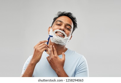 Grooming And People Concept - Young Indian Man Shaving Beard With Manual Razor Blade Over Grey Background