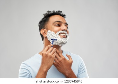 Grooming And People Concept - Young Indian Man Shaving Beard With Manual Razor Blade Over Grey Background