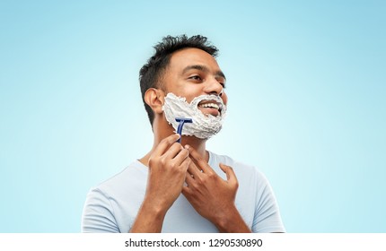 Grooming And People Concept - Young Indian Man Shaving Beard With Manual Razor Blade Over Blue Background