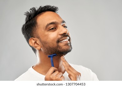 Grooming And People Concept - Young Indian Man Shaving Beard With Manual Razor Blade Over Grey Background