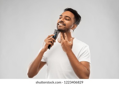 Grooming, Technology And People Concept - Smiling Indian Man Shaving Beard With Trimmer Over Grey Background