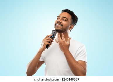 Grooming, Technology And People Concept - Smiling Indian Man Shaving Beard With Trimmer Over Blue Background