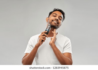 Grooming, Technology And People Concept - Smiling Indian Man Shaving Beard With Trimmer Over Grey Background