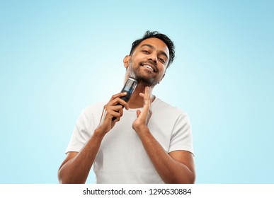 Grooming, Technology And People Concept - Smiling Indian Man Shaving Beard With Trimmer Over Blue Background