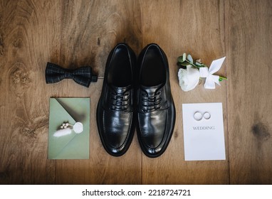 Groom's black shoes, bow tie, boutonniere, gold rings, an envelope with a heart stamp, a sheet of paper lie on a wooden background. Wedding photo of accessories and details, top view.