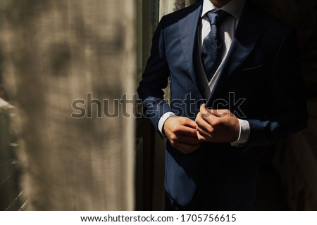 Grooms morning preparation. Cropped photo of elegant young fashion man dressing up for wedding celebration. Groom dressed in modern blue formal suit, white shirt getting ready for event. Sunny morning
