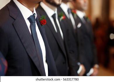 Groomsmen In Black Suits With Red Boutonnieres Stand In The Row