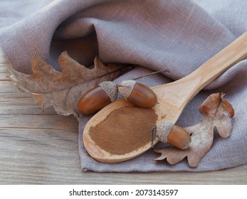 Ground acorn powder in wooden spoon, oak fruits, acorns and  autumn yellow leaves, napkin on a wooden table, closeup, flat layout. Natural decaffeinated coffee for use in dietary nutrition