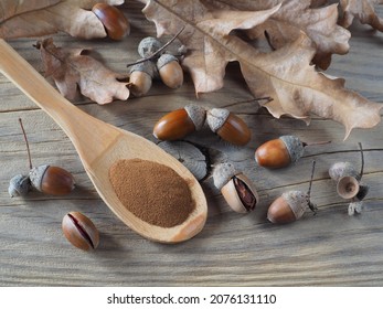 Ground acorn powder in wooden spoon, oak fruits, acorns and leaves on a wooden table, top view, flat layout. Natural decaffeinated coffee for use in dietary nutrition