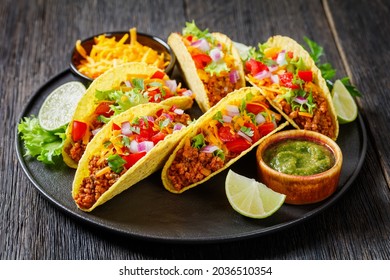 Ground Beef Tacos with shredded cheese, green salsa verde sauce, fresh lettuce, tomato, onion on a black plate with lime - Powered by Shutterstock