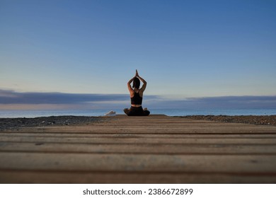 Ground level back view of anonymous female sitting in lotus pose with namaste hands above head while practicing yoga on seashore near sea