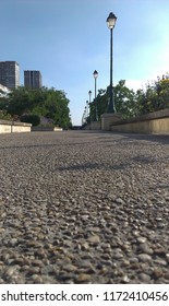 Ground Level Shot Of Concrete Pathway In Paris With Old Street Lamps On One Side. Pebbly Surface On Concrete Street