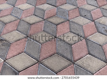 Ground level view of interlocking paving blocks in a trompe l'oeil (deceptive) design, resembling three-dimensional boxes. The material is rough, colored cement suitable for driveways or patios.