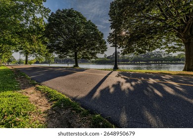 A ground level view of a paved walking path in Arthur J. Hendrickson Park in Valley Stream, Long Island in NY. The green trees cast shadows onto the paved path. A still lake is in the background.