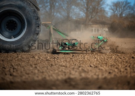 Ground level view shows a tractor tire and green cultivator tear dry soil, dust rising as metal tines churn clods. Leafless trees and warm afternoon light suggest spring.