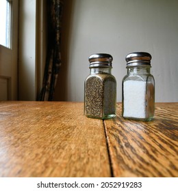 Ground Pepper And Salt Shakers On A Polished Wood Surface With Light From A Window Illuminating The Restaurant Table; Spices For Adding Flavor And Taste To Bland Food.