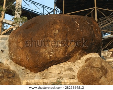 A grounded perspective highlighting a single colossal brown rock boulder prominently positioned on a masonry wall, set against a contrasting background of metal beams and a wooden deck.