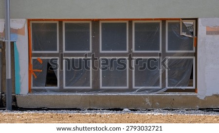 A ground-level ten-pane window structure is taped with orange adhesive and clear protective plastic film on an unfinished exterior wall above a dirt ground