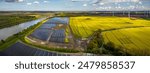 Ground-mounted solar photovoltaic panels, wind turbine farm neighboring canal, where ship gracefully navigates waterway. Photovoltaic park, windmill park, rapeseed field freight ship on Kiel Canal. 