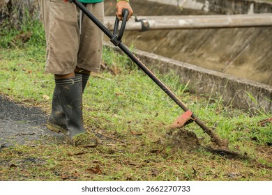 A groundskeeper in rubber boots uses a string trimmer to cut grass. A dynamic action shot of professional lawn care and landscape maintenance.
