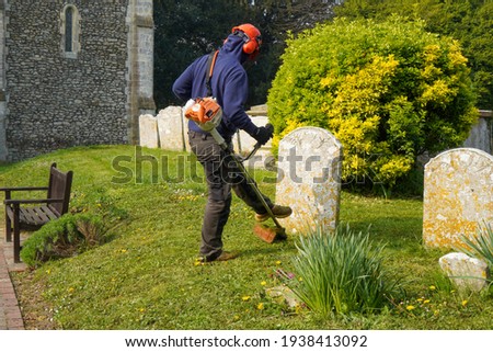Groundsman wearing an orange safety helmet and ear defenders at work in a local churchyard cutting grass around stone memorial gravestones with a petrol strimmer. The ancient Sussex flint church 