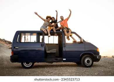 A group of 3 millennial mulitetnicos are having fun sitting on the roof of a camper van playing a flamenco guitar. Young people wave with their arms in the air. Spend the summer traveling with a van. - Powered by Shutterstock