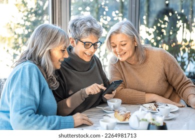 Group of 3 smiling mature female friends in white hats are using smartphone while at cafe having breakfast . Three senior women laugh look at the phone together - Powered by Shutterstock