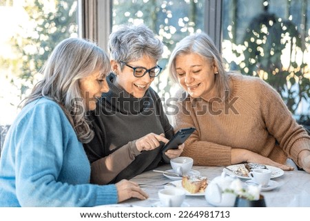 Group of 3 smiling mature female friends in white hats are using smartphone while at cafe having breakfast . Three senior women laugh look at the phone together