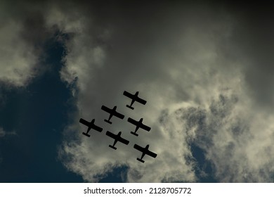 Group Of 6 Military Planes In Triangle Formation, Flying Overhead In The Sky. Camera Angle Looking Up. Cloudy Sky. Plane Is A Black Silhouette Shape. Concept For War. Planes On The Bottom Right.