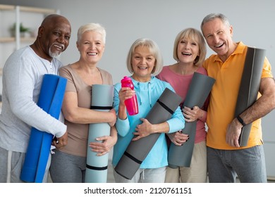 Group of active athletic pensioneers different nationalities in sportrswear attending yoga class at retreat center, holding fitness mats and cheerfully smiling at camera. Sport for senior people