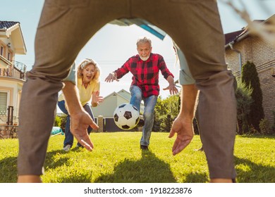 Group of active elderly people having fun playing football on the lawn in the backyard, enjoying sunny summer day outdoors, performing a free kick