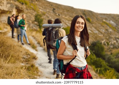 Group of active friends hiking or trekking on mountain trail on warm autumn weekend. Happy young woman hiker standing on hill pathway with blurry landscape in background, looking at camera and smiling - Powered by Shutterstock