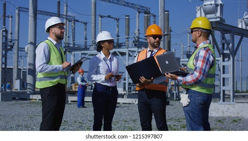 Group of adult diverse men in hardhats with gadgets gathering on transformer platform of solar electrical plant having discussion