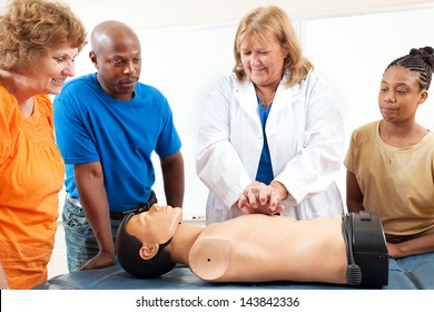 A Group Of Adult Education Students Watch A Doctor Or Nurse Demonstrating CPR Chest Compressioon On A Dummy.