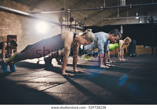 Group of adults doing push up exercises at indoor\
physical fitness cross-training exercise facility with bright light\
flare over them