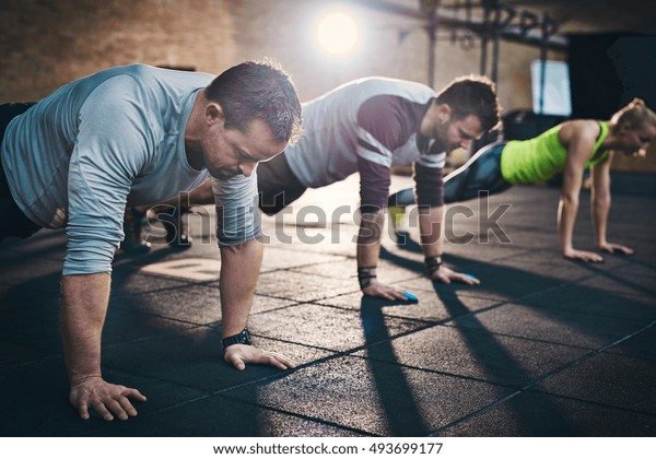 Group of adults performing push up exercise\
drills at indoor physical fitness cross-training exercise facility\
with bright light flare over\
them
