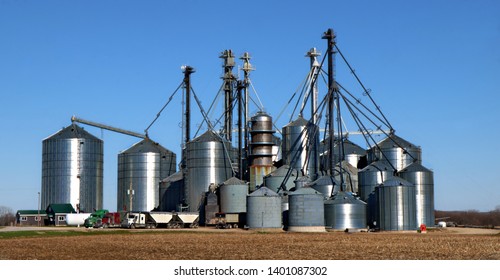 Group Of Agricultural Metal Grain Storage Bins With Dryer And Trucks Loading And Unloading. Business Trading Farm Commodities Concept