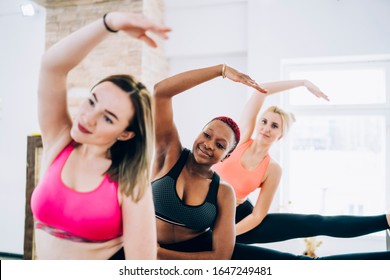 Group of amateur smiling multiethnic sportswomen standing with one leg on barre and reaching over leg with opposite arm in bright fitness room