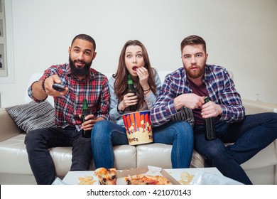 Group Of Amazed Shocked Young Friends Watching Tv And Eating Popcorn On Sofa