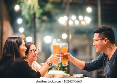 Group Of Asian Friends Or Coworkers Cheering With Beer, Celebrating Together At Restaurant Or Night Club. Young People Toasting At Party Event After Work. Success Or Friendship Concept. Focus On Glass