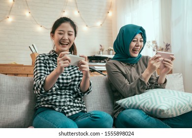 Group Of Asian Girl Friends Having Fun At Dining Room And Looking At Smart Phone. Chinese And Muslim Women Roommates Watching Football Game In Cellphone. Ladies Sisters Relax On Couch In Cozy House.