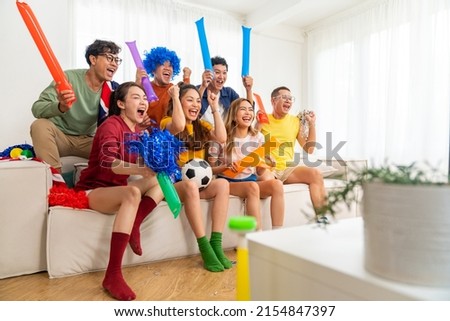 Group of Asian people friends sit on sofa watching and cheering soccer games competition on TV together at home. Happy man and woman sport fans shouting and celebrating sport team victory sports match