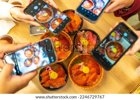 Group of Asian people using mobile phone photography seafood sashimi rice bowl in restaurant during travel fish market in Tokyo city, Japan. Tourist enjoy eating Japanese food donburi at street market