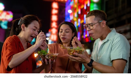 Group of Asian woman and LGBTQ people friends tourist enjoy eating traditional street food bbq seafood grilled squid with spicy sauce together at china town street night market in Bangkok, Thailand