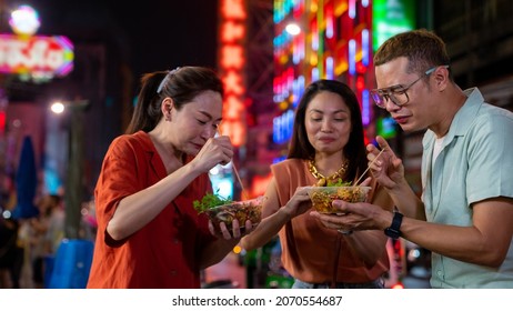 Group Of Asian Woman And LGBTQ People Friends Tourist Enjoy Eating Traditional Street Food Bbq Seafood Grilled Squid With Spicy Sauce Together At China Town Street Night Market In Bangkok, Thailand