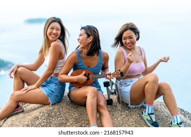 Group Of Asian Woman Sitting On Mountain Peak Playing Ukulele And Dancing Together At Tropical Island On Summer Travel Vacation. Female Friends Enjoy Outdoor Lifestyle Hiking And Skating Together