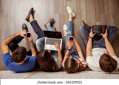 Group of attractive young people sitting on the floor using a laptop, Tablet PC, smart phones, headphones listening to music, smiling