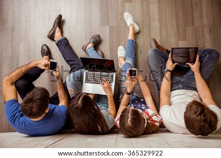 Group of attractive young people sitting on the floor using a laptop, Tablet PC, smart phones, headphones listening to music, smiling