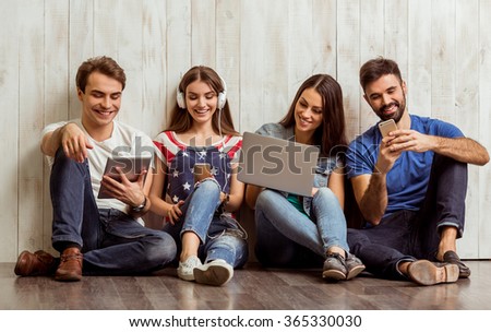 Group of attractive young people sitting on the floor using a laptop, Tablet PC, smart phones, headphones listening to music, smiling
